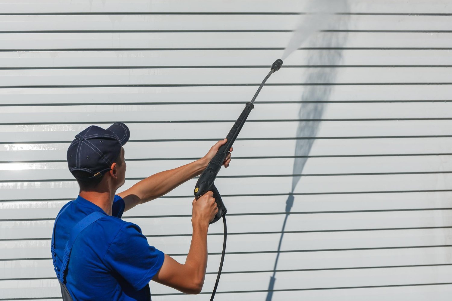 Person in a blue shirt and cap uses a pressure washer to clean a white siding wall, spraying water outward from the wand.