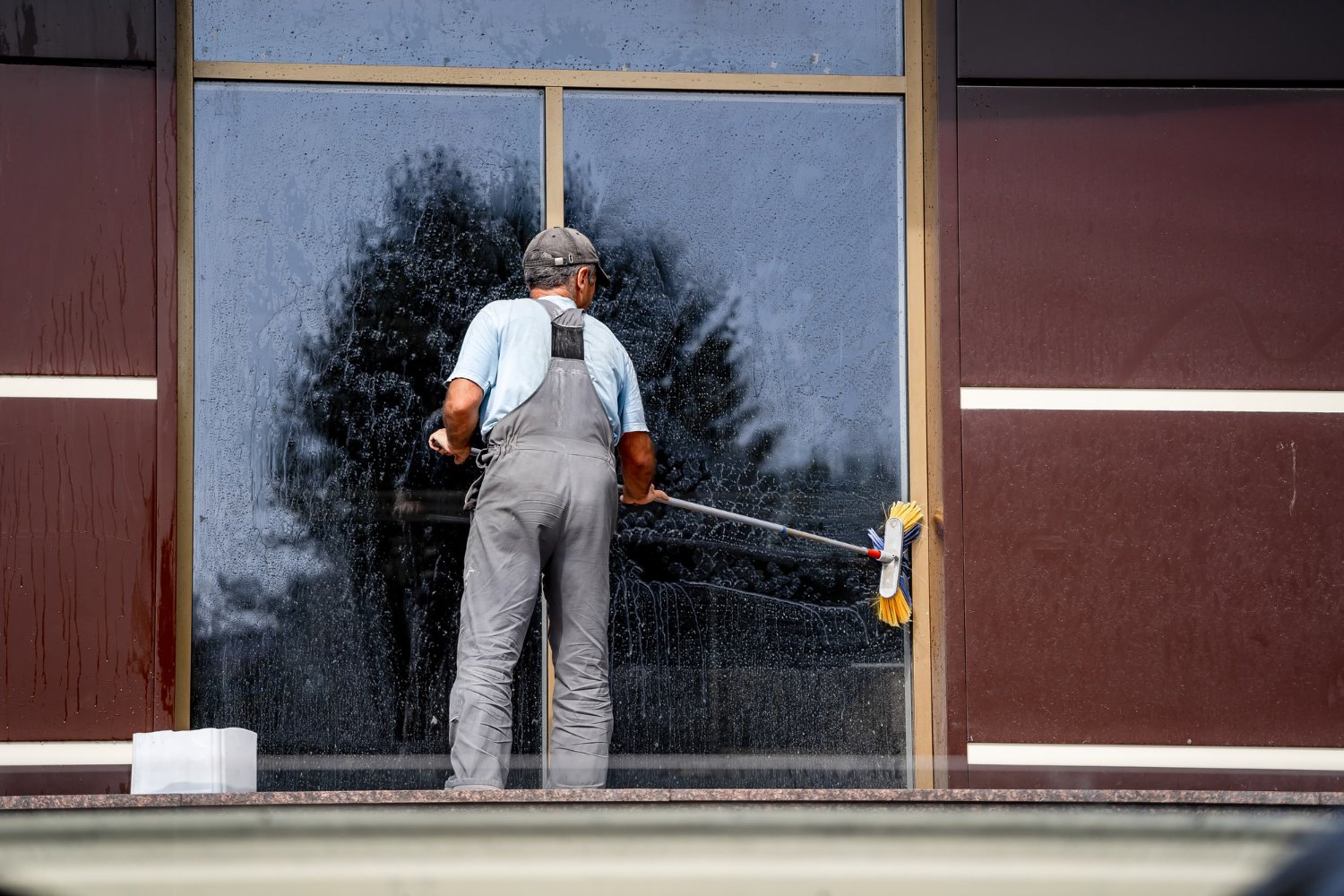 Window cleaner in gray overalls washing a large glass storefront with a long-handled brush outside the building